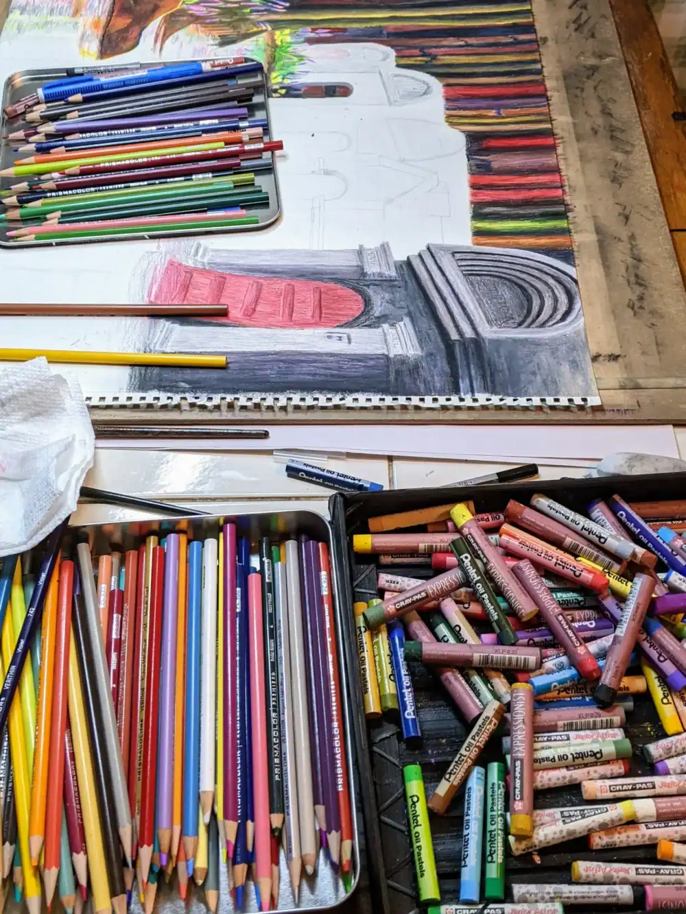 Colorful colored pencils and pastel sticks arranged around a partially completed architectural painting, showcasing artistic tools and creative process.