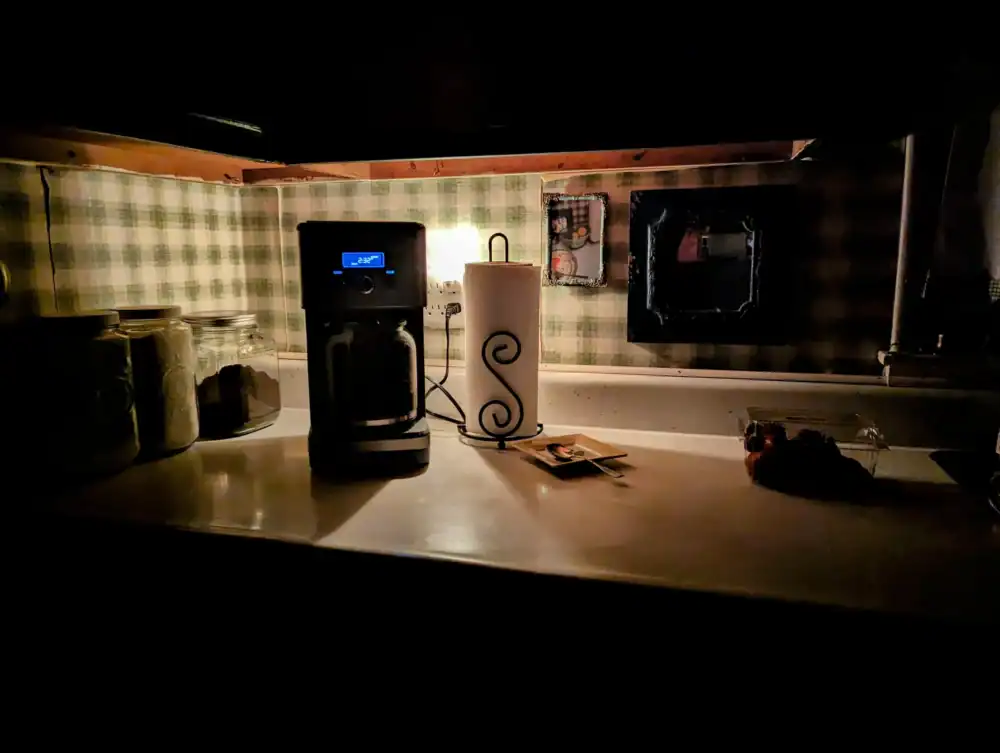A dimly lit kitchen counter with a glowing coffee maker, jars, and a paper towel holder against checkered green and white wallpaper, capturing a warm, rustic ambiance.