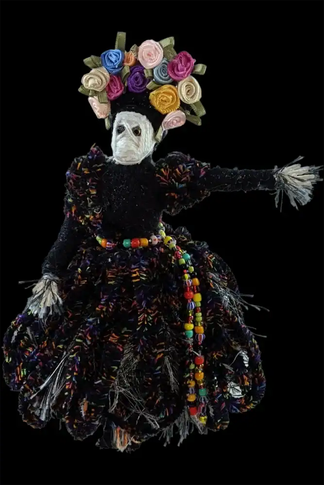 Close-up of a handcrafted female Day of the Dead skeleton doll wearing a colorful floral headdress and dark, but also colorful, bead-belted dress.
