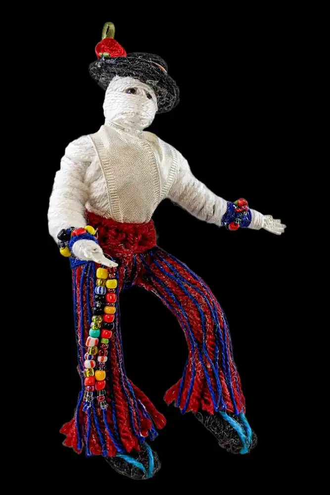 Front view of a handcrafted male Day of the Dead skeleton doll, wearing a black hat with a red rose, a white shirt, and colorful pants with a beaded belt.