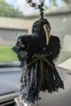 Side angle of a Plague Doctor Charm showing the detailed mask, button face, and knotted yarn cloak hanging from a car's rearview mirror.