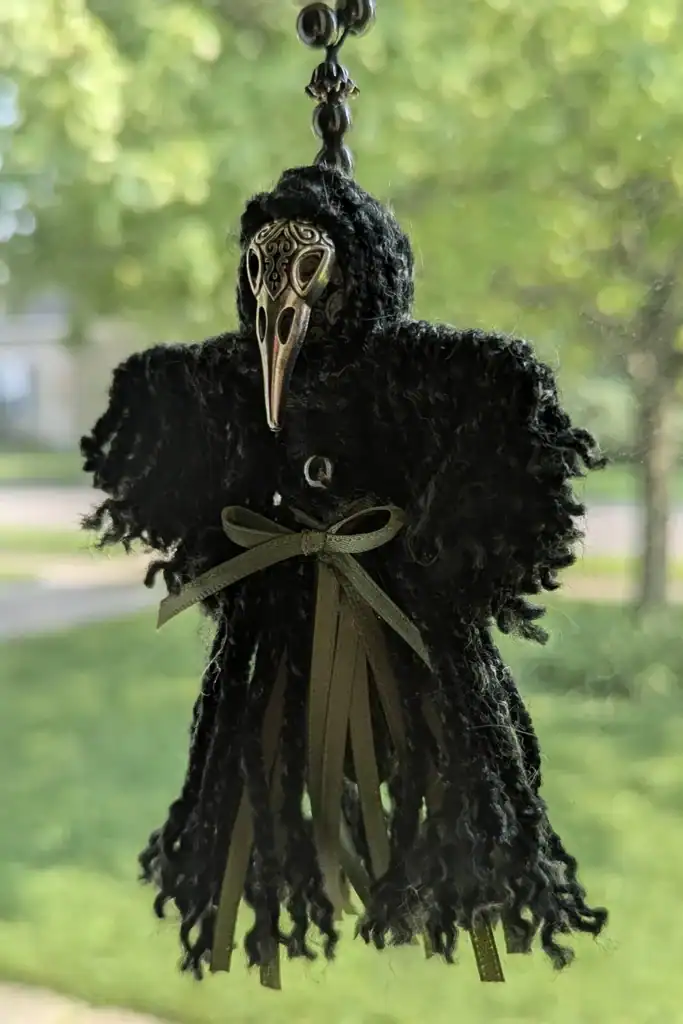Left front angle of a Plague Doctor Charm with a metal mask and green ribbon, hanging in a window.