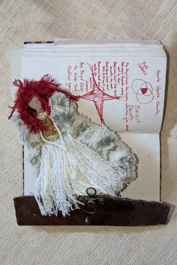 Brigit You-Do-You Voodoo™ doll resting on an open journal page showing red ink drawings and handwritten prayers, including Brigid’s cross, triple goddess, and symbolic notes.