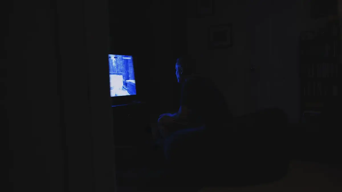 A man sits on a bean bag chair in a dim living room at night, lit only by a television screen, seen from a hallway doorway.